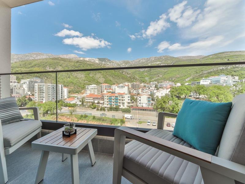 Balkon mit Stühlen und kleinem Tisch, Blick auf Wohnhäuser und grüne Hügel unter blauem Himmel.