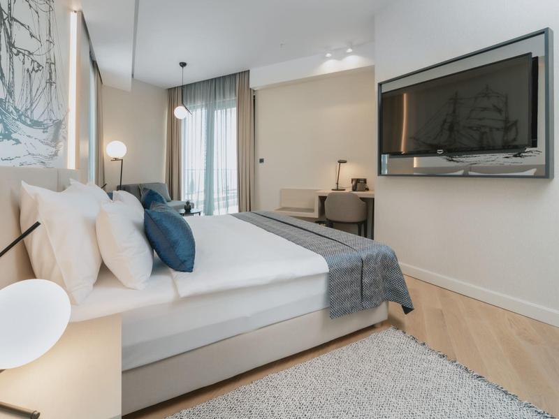 Modernes Schlafzimmer mit großem Bett, weißen Wänden, dunklen Kissen und hellgrauem Teppich.