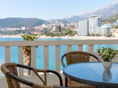 Balkon mit Tisch, zwei Stühlen und Vase, Blick auf Meer, Strand, Palmen und Häuser am Berg.