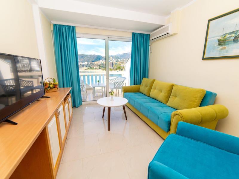 Helles Wohnzimmer mit blau-grüner Couch, TV auf Sideboard und Blick auf Balkon mit Meerblick.
