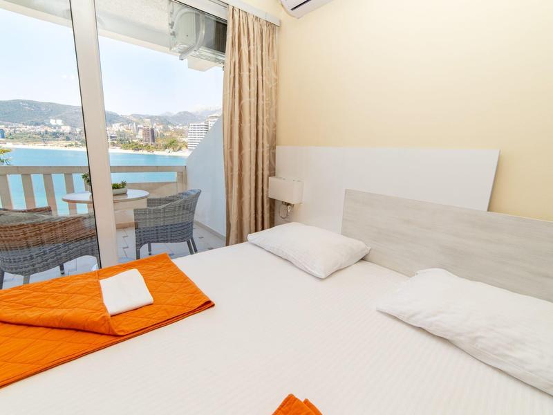 Helles Schlafzimmer mit großem Bett, orangefarbener Decke und Balkon mit Blick auf Wasser und Berge.