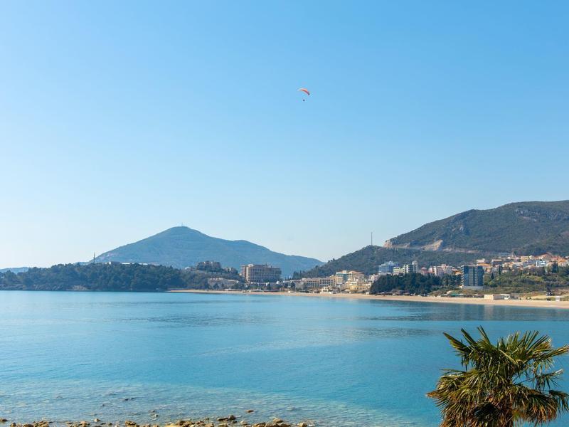 Klares Meer vor Küste mit Gebäuden, Bergen im Hintergrund und blauem Himmel.