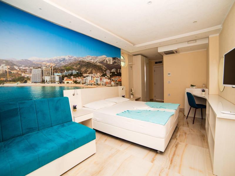 Modern eingerichtetes Zimmer mit weißem Bett, türkisfarbenem Sofa und Wandbild einer Küstenlandschaft.