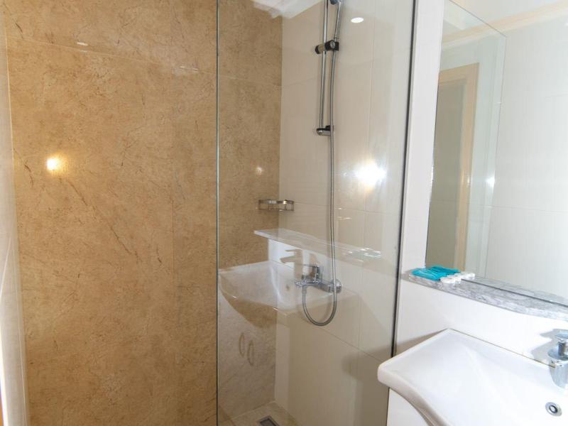 Kleines Badezimmer mit Glasdusche, weißem Waschbecken und Spiegel, beige Fliesen an Wand und Boden.