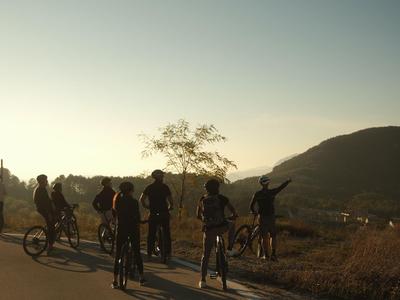 Groep fietsers op een landweg bij zonsondergang met heuvels op de achtergrond.