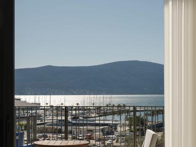 Blick durch eine Tür auf einen Balkon mit Stühlen und einem Tisch, dahinter ein Hafen und Berge.