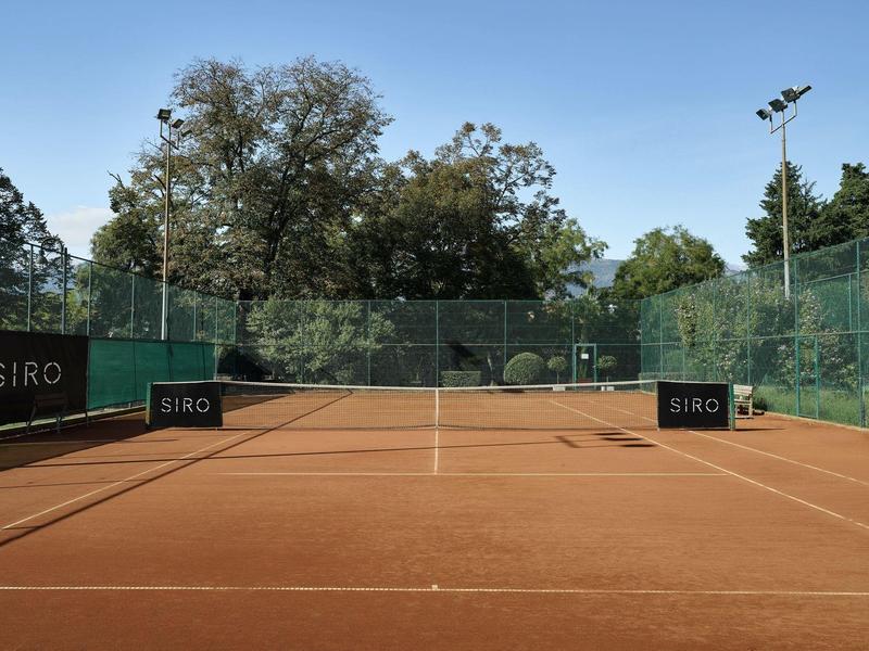 Rotes Tennisplatz mit Bäumen und Zäunen bei sonnigem Wetter.