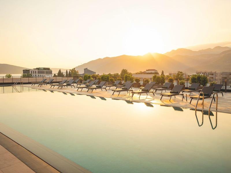 Großer Pool mit Liegen bei Sonnenuntergang, umgeben von Bergen und Hotelgebäuden.