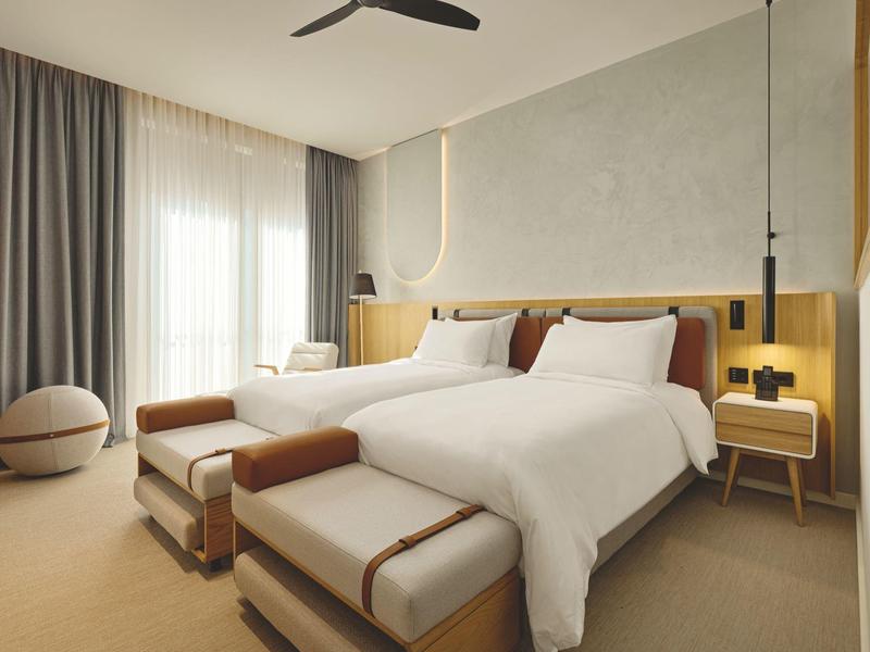 Modernes Hotelzimmer mit zwei Doppelbetten, Sitzbänken und großen Fenstern mit Vorhängen.