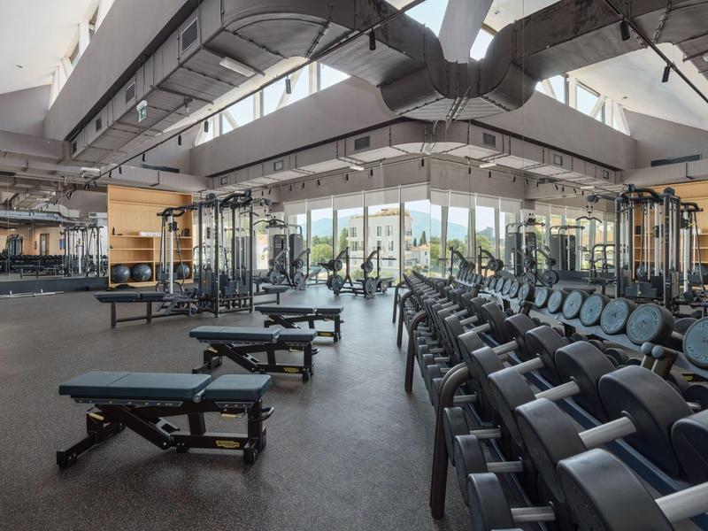 Geräumiger Fitnessraum mit Gewichten, Bänken und großen Fenstern mit Blick ins Freie.