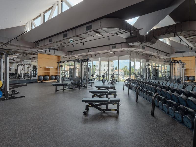 Großer Fitnessraum mit Gewichten, Bänken und viel natürlichem Licht durch große Fenster.