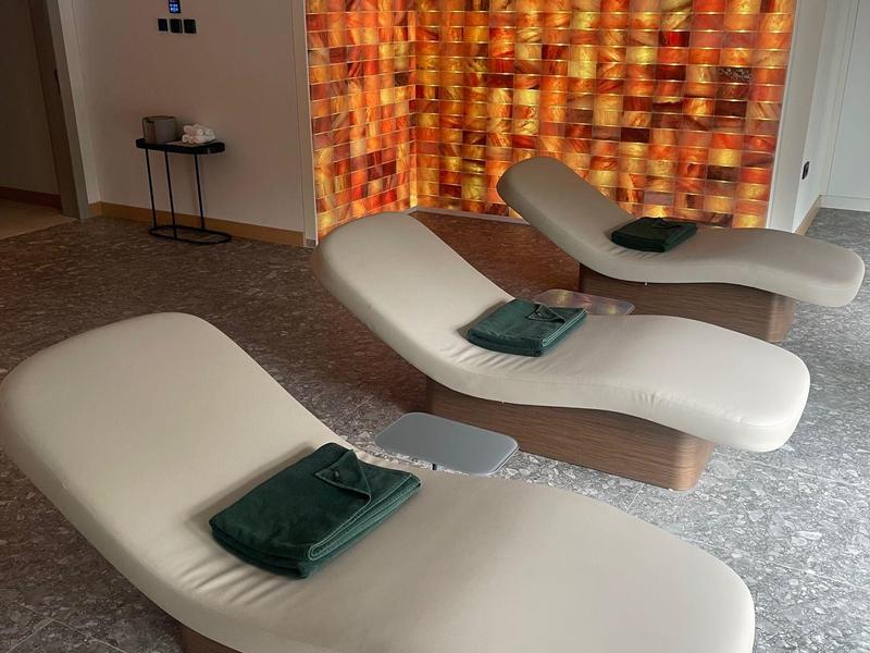 Drei moderne Liegen mit Handtüchern in einem ruhigen Wellnessraum mit warm beleuchtetem Steinmosaik.