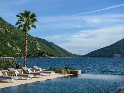 Piscine à débordement avec chaises longues près de palmiers et montagnes au bord de la mer sous un ciel bleu.