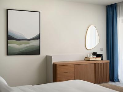 Helles Hotelzimmer mit modernem Sideboard, abstraktem Bild und dreieckigem Spiegel an der Wand.