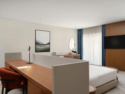 Modernes Hotelzimmer mit Doppelbett, Schreibtisch, Stuhl, TV und Fenster mit Vorhängen.