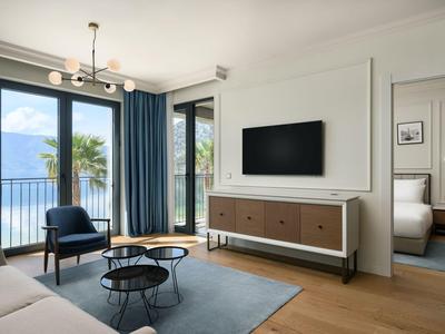 Modernes Wohnzimmer mit Blick auf einen See, zwei Stühlen, Fernseher und großem Fenster mit Vorhängen.