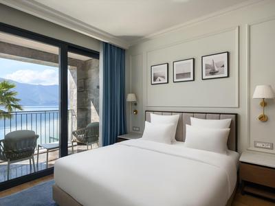 Helles Hotelzimmer mit großem Bett und Balkon mit Blick auf See und Berge.