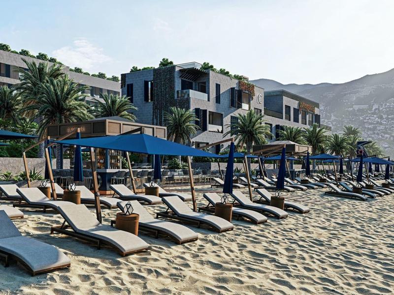 Strand mit Liegestühlen und blauen Sonnenschirmen vor einer Hotelanlage und Bergen im Hintergrund.