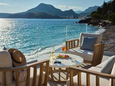 Terrasse mit Holzmöbeln, Blick auf Meer und Berge bei sonnigem Wetter.