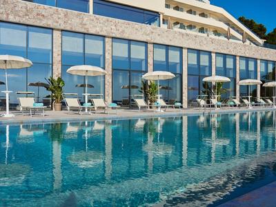 Nowoczesny basen hotelowy z leżakami i parasolami przed szklaną elewacją.