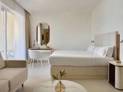 Jasny pokój hotelowy z podwójnym łóżkiem, biurkiem, krzesłem i sofą przy balkonie.