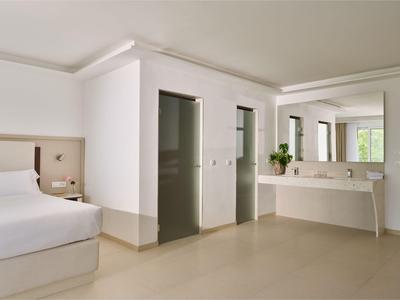 Nowoczesny pokój hotelowy z białym łóżkiem, dużym lustrem i otwartymi drzwiami do dwóch łazienek.