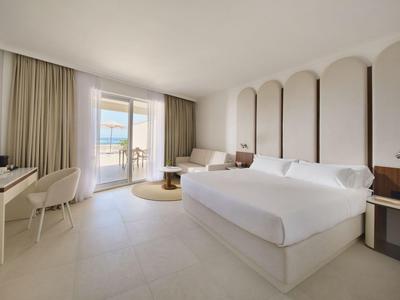 Jasny pokój hotelowy z łóżkiem king-size, fotelami i balkonem z widokiem na morze.