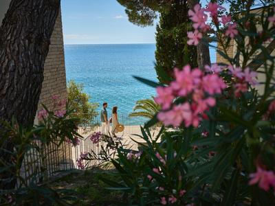 Blick auf ein Meer mit einem Paar, das am Strand neben Blumen in einem Hotelgarten steht.