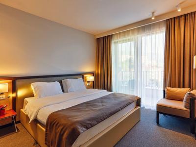 Helles Hotelzimmer mit Doppelbett, Nachttischen, Sessel und großen Fenstern mit Vorhängen.