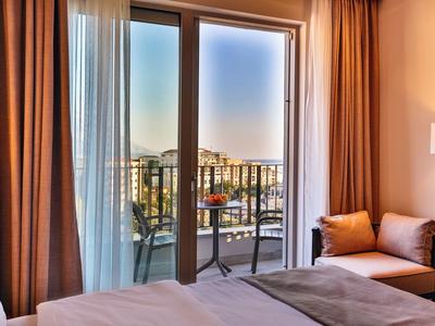 Helles Hotelzimmer mit Balkon, Tisch und Stühlen mit Ausblick auf die Stadt bei Sonnenuntergang.