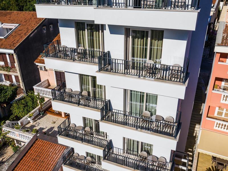 Außenansicht eines modernen weißen Gebäudes mit mehreren Balkonen und Möbeln in einer städtischen Umgebung.