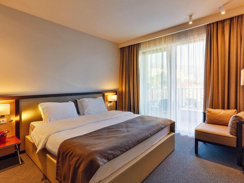 Helles Hotelzimmer mit Doppelbett, Nachttischen, Sessel und großen Fenstern mit Vorhängen.