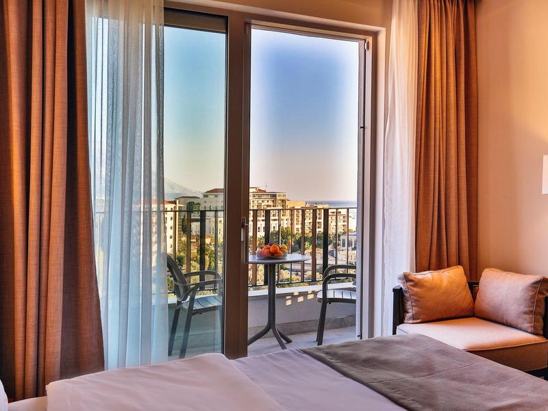 Helles Hotelzimmer mit Balkon, Tisch und Stühlen mit Ausblick auf die Stadt bei Sonnenuntergang.