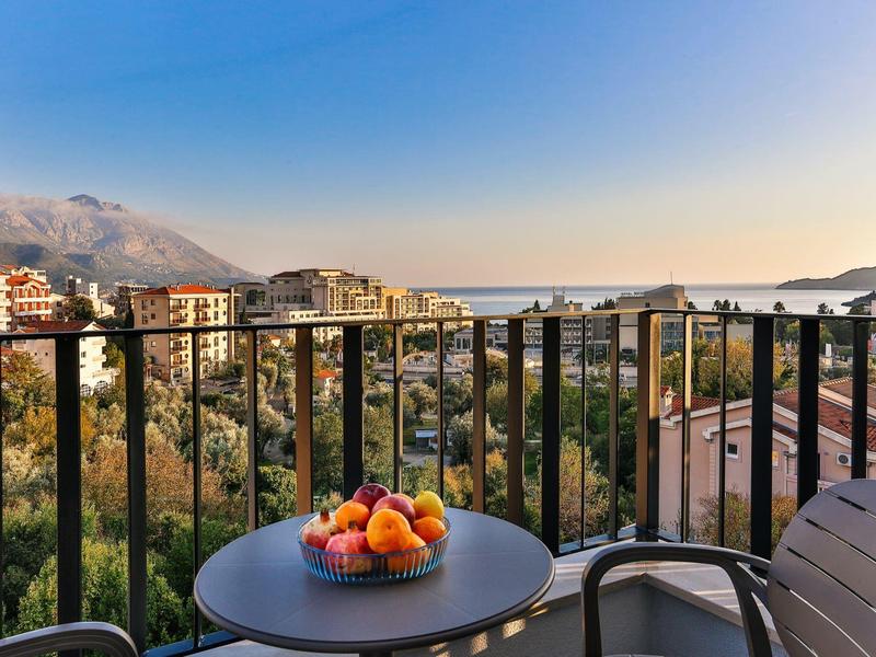 Balkon mit Tisch und Obstschale, Blick auf Häuser und Berge bei klarem Himmel.