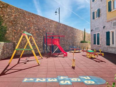 Kinderspielplatz mit Rutsche, Schaukeln und Wippe auf rotem Boden neben einem Gebäude und einer Steinmauer.