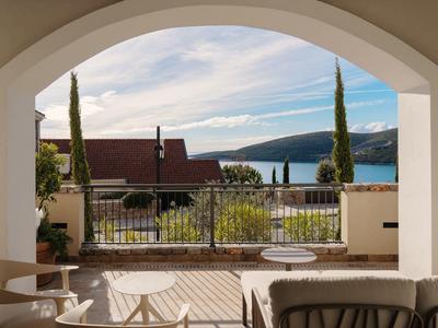Blick von einer überdachten Terrasse auf einen Garten mit Meer und Bergen im Hintergrund.