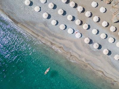 Luftaufnahme eines Strandes mit Reihen von weißen Sonnenschirmen und einem Paddelbrett im Wasser.