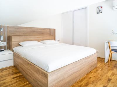 Helles Hotelzimmer mit Holzbett, weißer Bettwäsche, Nachttisch und Stuhl am Fenster.