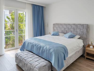 Helles Schlafzimmer mit grauem Bett, blauer Bettwäsche und Balkon mit Blick auf grüne Bäume.