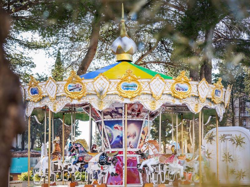 Bunter Karussell mit Pferden in einem Park, umgeben von Bäumen und grüner Natur.