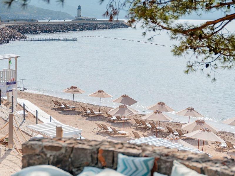 Strand mit Sonnenschirmen und Sitzbank mit Kissen unter einem Baum am Wasser