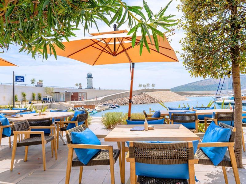 Outdoor-Café mit Holztischen, blauen Kissen und orangefarbenen Sonnenschirmen am Wasser.