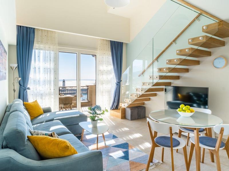 Hell eingerichtetes Wohnzimmer mit blauen Vorhängen, grauem Sofa, Treppe und Essbereich mit Holzmöbeln.