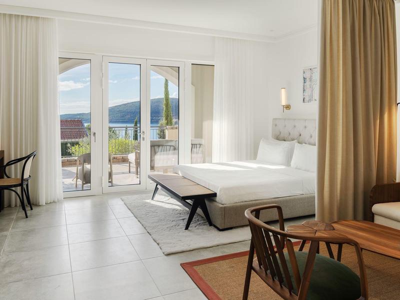 Helles Hotelzimmer mit großem Bett, Schreibtisch, Sessel und Blick auf Terrasse und Berge.