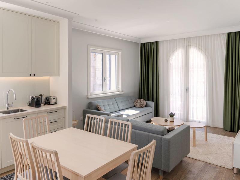 Helle Hotelwohnung mit Küchenzeile, Esstisch, Sofa und großen Fenstern mit grünen Vorhängen