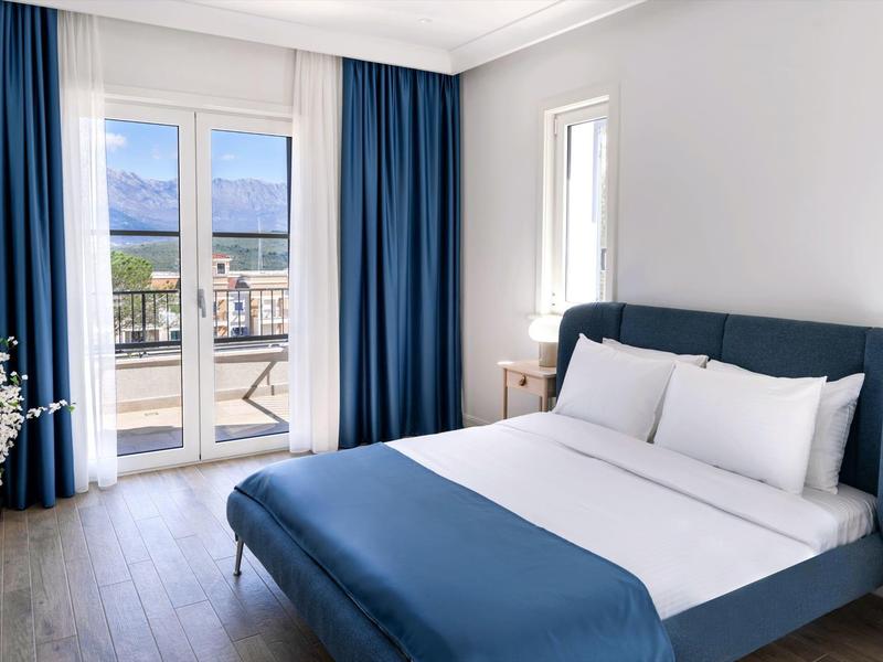 Modernes Gästezimmer mit großem Bett, blauen Vorhängen und Zugang zu Balkon mit Blick auf Außenbereich.