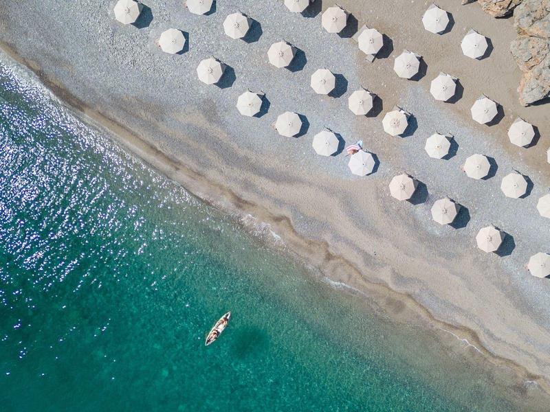 Luftaufnahme eines Strandes mit blauem Wasser und Reihen von weißen Sonnenschirmen am Sandstrand.