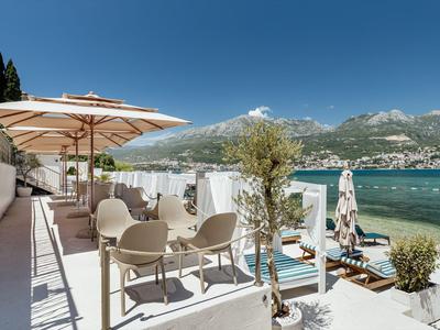Terrazza con tavoli, sedie e sdraio con vista sul mare e le montagne sotto un cielo sereno.