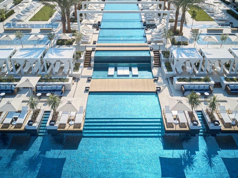 Großer moderner Pool mit mehreren Wasserbecken, Liegen und weißen Sonnenschirmen, umgeben von Gebäuden.