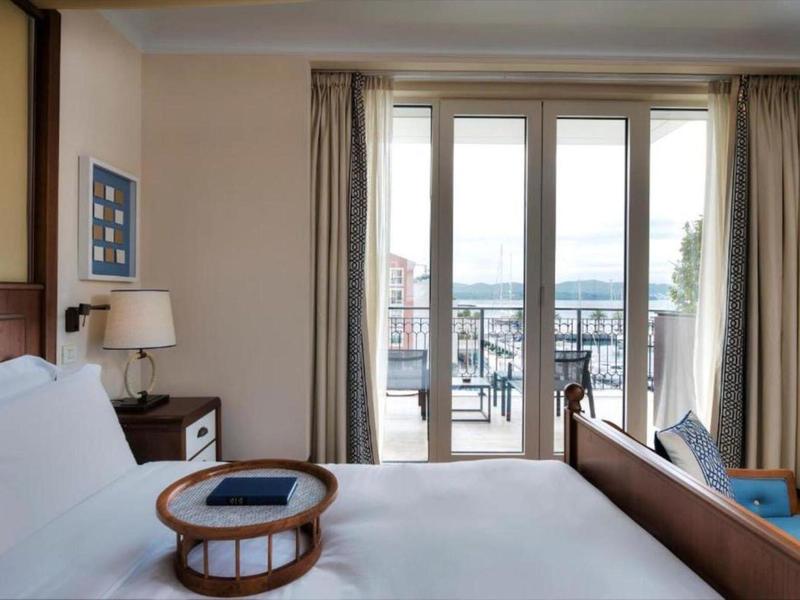 Helles Schlafzimmer mit weißen Wänden, Bett, Nachttisch, Lampe und Balkon mit Glas-Schiebetür.